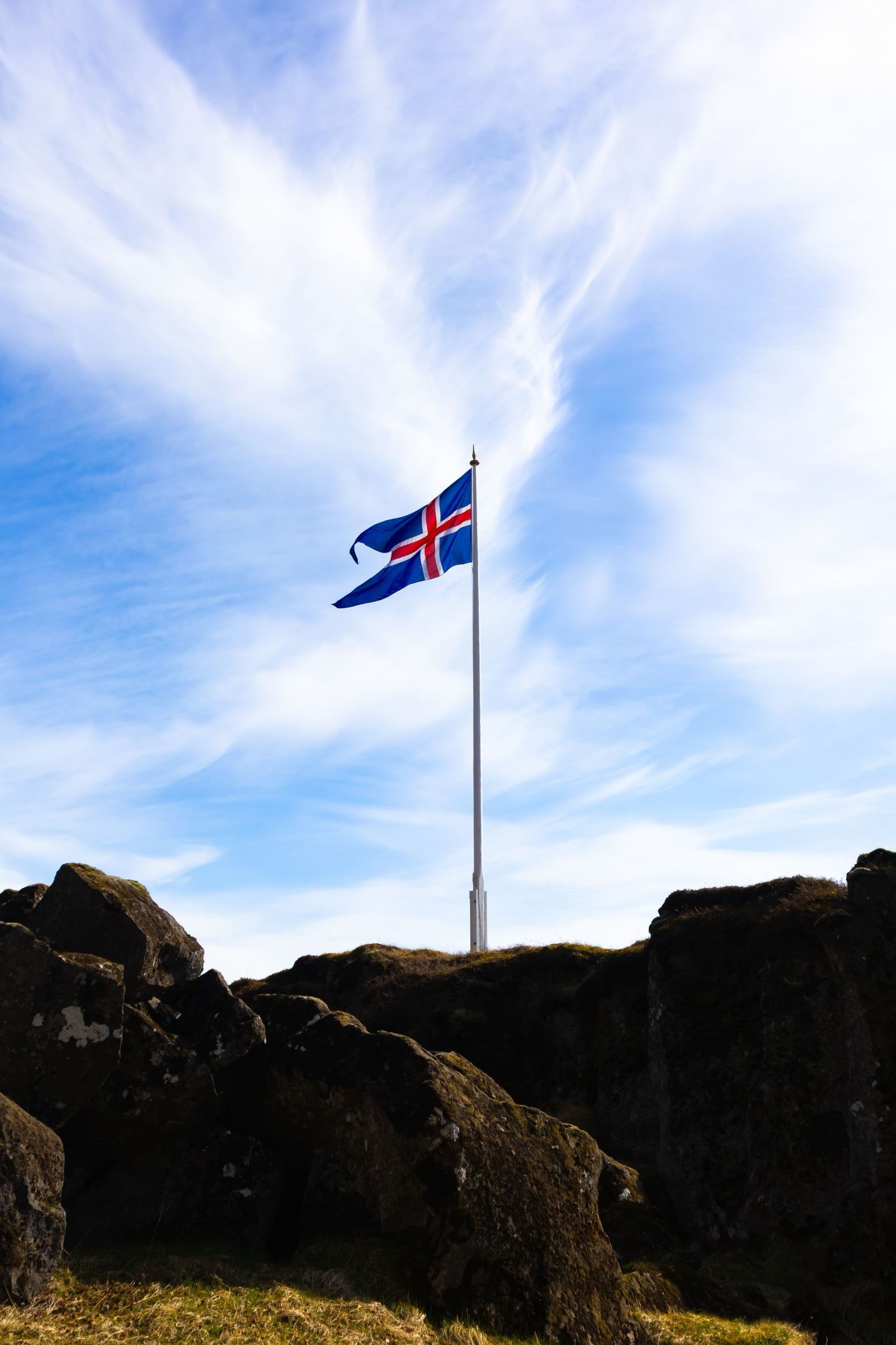 The Icelandic flag flying above dark rocks under a wide blue sky