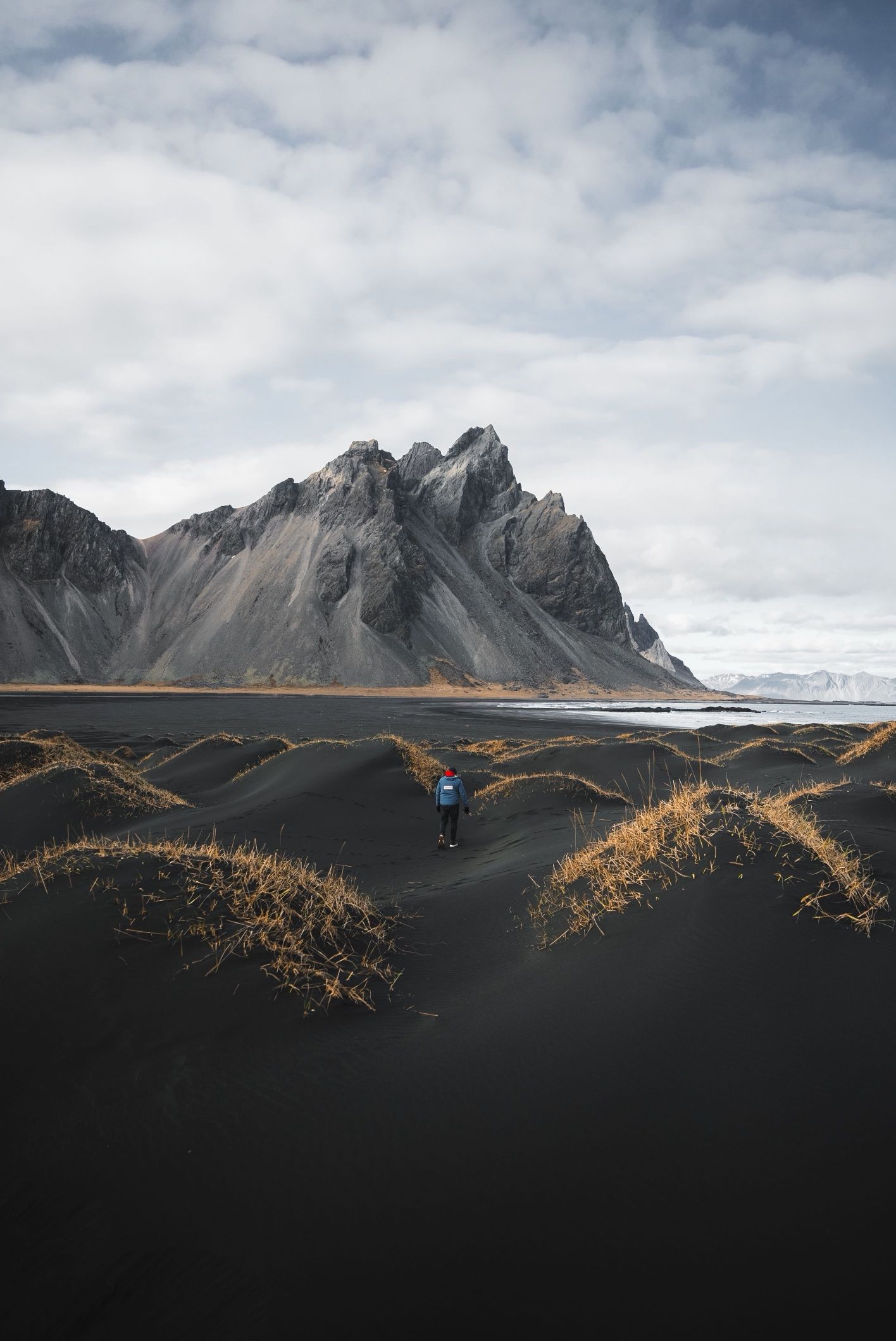 A walker in a blue jacket crossing grass-tufted black dunes below the jagged peaks of Vestrahorn