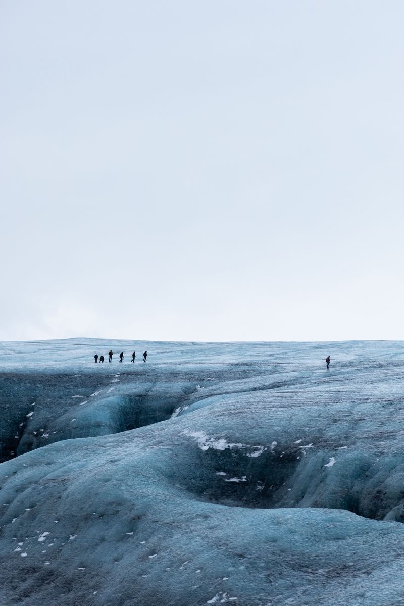 A line of hikers crossing a blue-grey glacier under a white sky