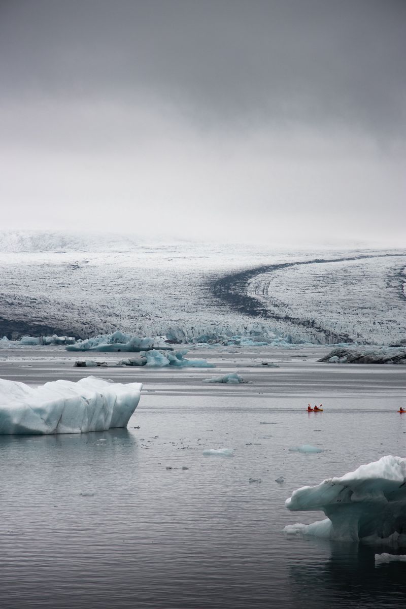 Two orange kayaks paddling between icebergs below a misty glacier