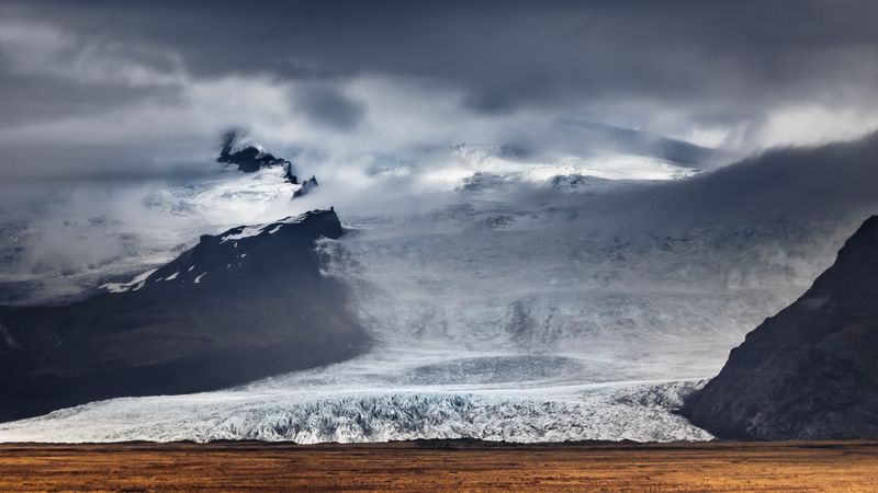A glacier spilling from cloud-covered mountains above golden grassland