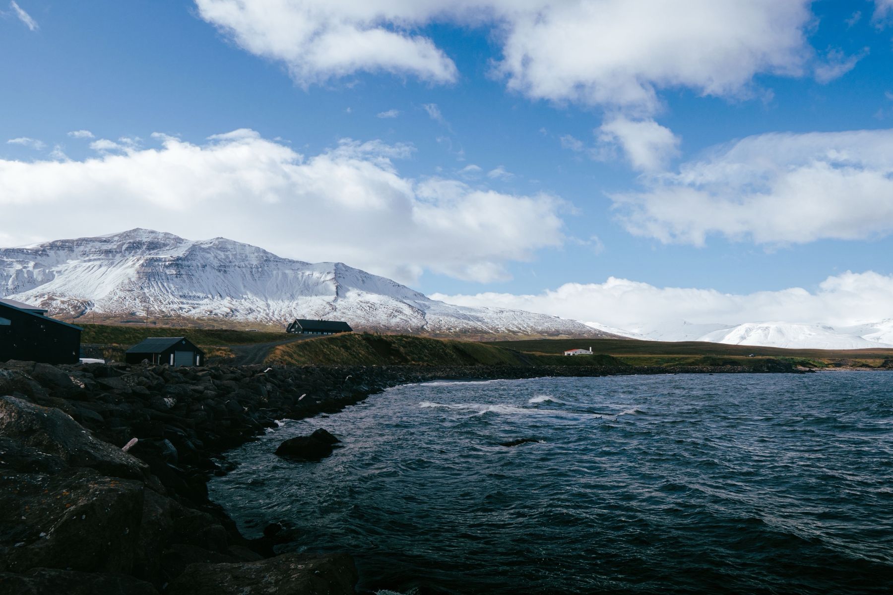 Snow-capped mountains above a rocky shore and dark wooden houses under a blue sky