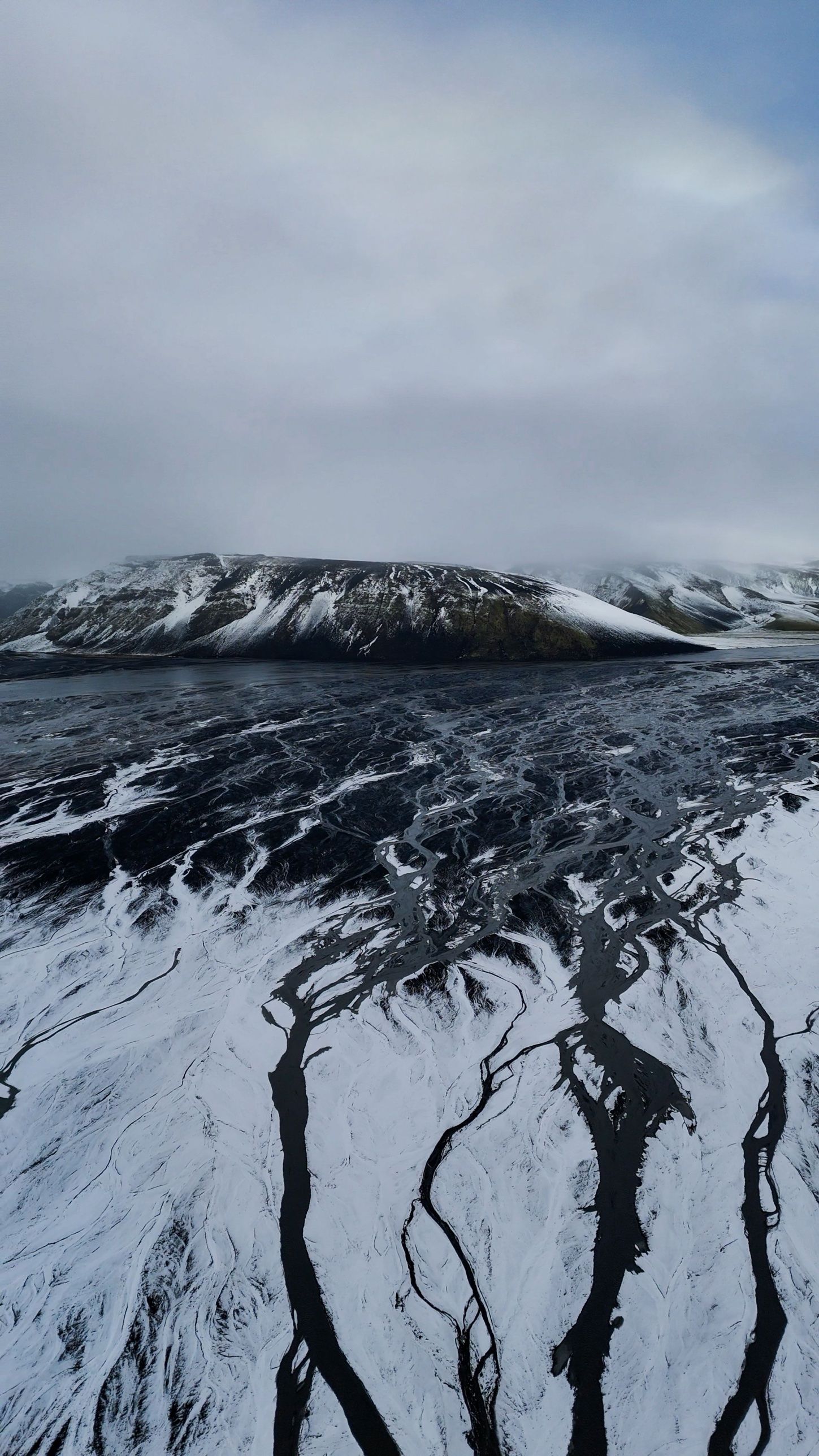 Black meltwater channels branching across a snow-covered plain below a dark mountain